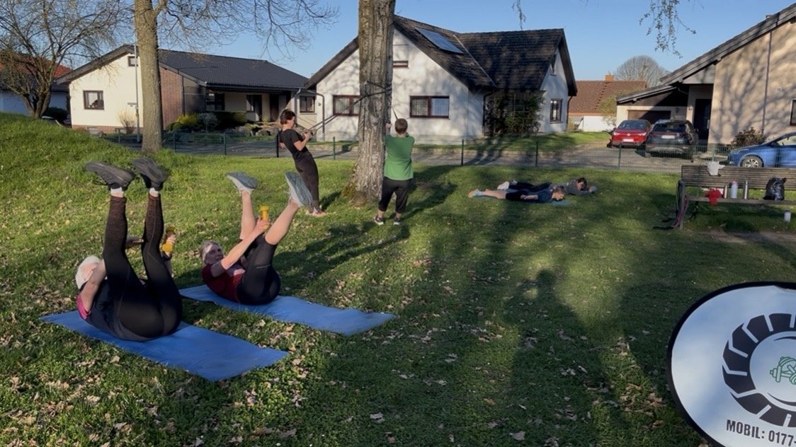 Teilnehmende liegen auf Matten im Garten und heben die Beine, BootKamp-Banner im Bild